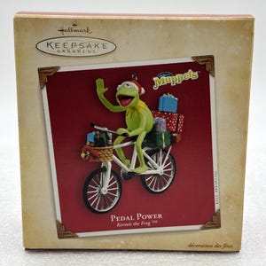 Puede incluir: Una caja de adorno Hallmark Keepsake con la rana Kermit montando una bicicleta blanca. Kermit lleva un gorro de Papá Noel y saluda, con regalos envueltos en una cesta delantera y en el portaequipajes. La caja muestra Pedal Power y Muppets.