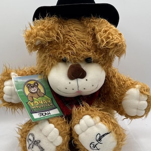 May include: A plush teddy bear named "Jean" with brown fur, a black hat, and a red plaid scarf. The bear has white paws with embroidered designs and a tag from the Yonah Mountain Collection.