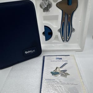 May include: A QuicKutz craft tool set featuring a blue and silver metal hand tool in a white foam tray, a dark blue zippered storage case with a QuicKutz logo patch, and a plastic-wrapped instruction guide detailing the included components.