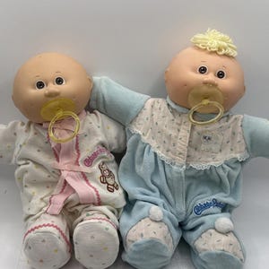 May include: Two Cabbage Patch Kids dolls. One doll wears a white and pink patterned outfit with a pink bow. The other doll wears a blue outfit. Both dolls have pacifiers and blonde hair.