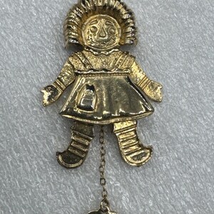 Puede incluir: Collar con colgante dorado con un diseño de muñeca estilizada. La muñeca tiene una cara, un vestido y piernas detallados. Un pequeño dije en forma de corazón con las palabras "I Love U" cuelga de los pies de la muñeca. La cadena del collar también es dorada.