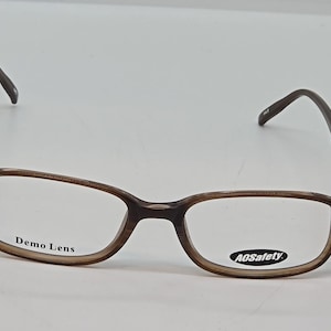 Puede incluir: Gafas con montura rectangular y patrón de carey marrón. El cristal izquierdo presenta el texto Demo Lens y el derecho muestra el texto AOSafety. Las gafas tienen patillas finas y curvadas y un puente que une ambos cristales.