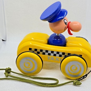 May include: A yellow wooden pull toy taxi with a black and white checkered stripe and the word TAXI printed on the side. A cartoon figure with a blue hat and a red mustache sits in the driver seat. The toy has four yellow wheels with silver centers and a green pull cord.