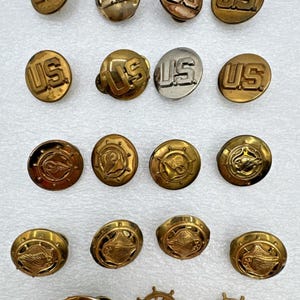 May include: A selection of antique brass buttons. Some feature the letters "US", while others display nautical designs. The buttons are in various sizes and finishes, reflecting different vintage styles.