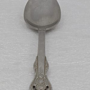 Può includere: Un cucchiaio color argento con una coppa ovale e un manico decorativo. Il manico presenta un design ornato con dettagli floreali e volute. Il cucchiaio è in metallo e probabilmente viene utilizzato per mangiare o servire cibo.