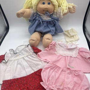 May include: A vintage Cabbage Patch Kid doll with blonde yarn pigtails, blue eyes, and a blue gingham dress. Includes a white dress with red trim, a pink dress and bloomers, and red floral bloomers.