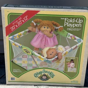 Può includere: Un box pieghevole per bambole Cabbage Patch Kids in una scatola con il testo Actual Size 20" x 20" x 12". La scatola mostra due bambole, una in piedi con un vestito rosa e una sdraiata, con un motivo a box verde e bianco sulla confezione.