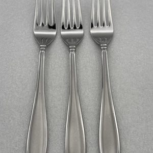 May include: Three polished stainless steel dinner forks arranged vertically. Each fork features four tines, a slender neck, and a flared handle with a central recessed panel bordered by raised lines, ending in a small decorative rounded tip.