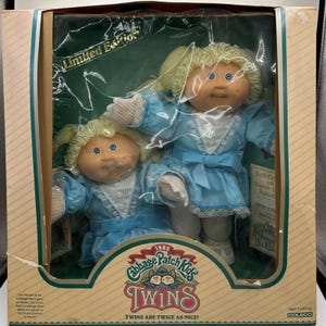 Puede incluir: Un juego de muñecas Cabbage Patch Kids Twins de 1985 en caja. Las muñecas tienen cabello rubio de hilo, ojos azules y visten vestidos azul claro a juego con ribete de encaje blanco. La caja dice "Edición Limitada" y "Twins are twice as nice!"