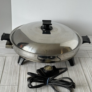 May include: A stainless steel electric skillet with a lid and black handles. The skillet sits on black legs and includes a temperature control and power cord. The lid has a black handle.