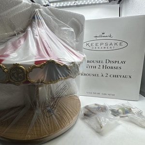 May include: A Hallmark Keepsake Ornament carousel display with two horses, featuring a red and white striped canopy and gold decorative trim. The set includes a tan circular base and two separate horse figures in plastic packaging.