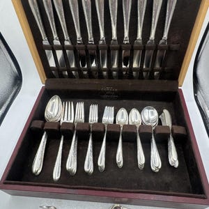 May include: A silver cutlery set housed in a wooden storage case with a dark brown velvet interior. The set features ten knives in the lid, and an assortment of forks and spoons in the base. Two extra pieces, a fork and a spoon, lie outside.