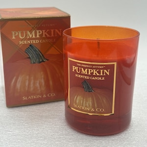 Puede incluir: Una vela aromática en un frasco de vidrio naranja y su caja de cartón a juego. Ambos presentan una etiqueta con el texto The Perfect Autumn Pumpkin Scented Candle Slatkin & Co. y un gráfico de una calabaza con un tallo verde oscuro.