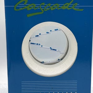 Puede incluir: Un temporizador de líquido rectangular azul Cascade Idroclessidra con un recorte circular en el centro. Dentro del círculo, un fondo blanco muestra estantes transparentes escalonados con pequeñas gotas de líquido azul.