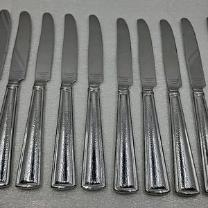 May include: A set of ten stainless steel dinner knives. Each knife has a long, slender blade and a textured handle with a flared, rectangular base. The knives are arranged in a row, reflecting light.