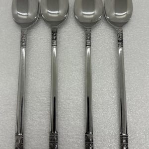 May include: Four silver spoons with elongated bowls and long, slender handles. The handles feature a decorative band near the bowl and a textured pattern at the base. The spoons are arranged in a row, reflecting light.