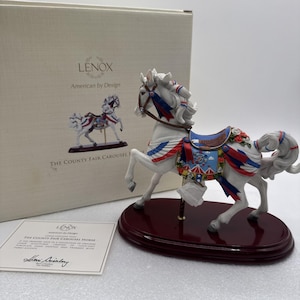 May include: A white porcelain carousel horse figurine featuring red, white, and blue ribbons, a saddle with a ferris wheel graphic, and red floral accents. The horse stands on a dark wood-toned oval base, accompanied by its original box and certificate.