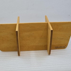 May include: A light brown wooden basket divider with a smooth finish and a trapezoidal shape. The piece features two vertical slots that intersect with a long horizontal panel. The text Longaberger Woodcrafts is printed on the lower right side.