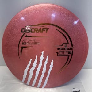 May include: A pink Discraft Undertaker distance driver disc with gold and white graphics. The disc features the text "Discraft", "Paul McBeth 5X World Champion", and "Undertaker Distance Driver". White claw marks are on the lower left.