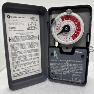 May include: A gray metal electrical timer with a red and white dial, displaying morning and evening hours. The timer has a black pointer and a warning label about electric shock. The model number is CMP-240.