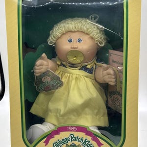 Pode incluir: Boneca Cabbage Patch Kids na caixa original. A boneca tem cabelo loiro de lã, olhos azuis e um vestido amarelo. A caixa apresenta o logotipo Cabbage Patch Kids e o ano de "1985". A boneca está usando uma chupeta.