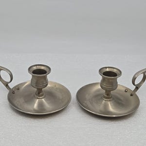 May include: A pair of silver-coloured candle holders. Each has a circular base, a central cup for the candle, and a curved handle. The candle holders are made of metal and have a simple, classic design. Suitable for home decor.