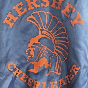 May include: A light blue jacket with orange lettering and a graphic. The word "HERSHEY" arches across the top, and "CHEERLEADER" curves along the bottom. An orange profile of a Native American chief is in the center.