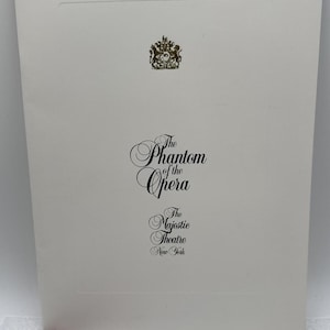 May include: A white rectangular programme booklet featuring a gold crest at the top and black text that reads The Phantom of the Opera, The Majestic Theatre, New York. A small red tassel is attached to the bottom left corner of the cover.