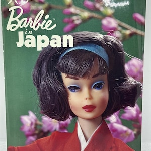 May include: A book cover featuring a Barbie doll in a red kimono with a blue headband. The title "Barbie in Japan" is displayed in white, with Japanese characters above. The background shows pink flowers and green foliage.