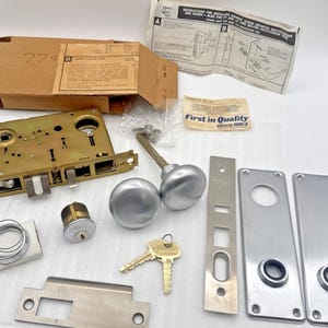 Könnte beinhalten: Ein Vintage-Türschloss-Set mit Messingmechanismus, silberfarbenen Türknöpfen und passenden Frontplatten. Enthält Schlüssel, einen Zylinderschloss und Anweisungen. Die Verpackung trägt die Aufschrift "First in Quality since 1964".