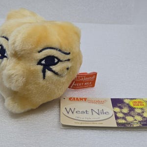 May include: A plush toy shaped like a virus, colored light yellow with embroidered dark blue eyes featuring an Eye of Horus design. An attached orange tag reads GIANTmicrobes WEST NILE, and a rectangular card displays the text West Nile.