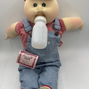 May include: A Cabbage Patch Kids doll, Ethan Prentice, wearing a red cap, red and white checkered shirt, and blue striped overalls. The doll is holding a white baby bottle and has red shoes. A tag is attached to the overalls.