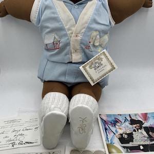Peut inclure: Poupée Cabbage Patch Kid vintage à la peau brune, vêtue d'une tenue bleu clair avec des bordures blanches et des chaussures assorties. La poupée a un visage rond et tient des papiers d'adoption et un certificat de naissance. Les chaussures de la poupée portent les initiales "X R".