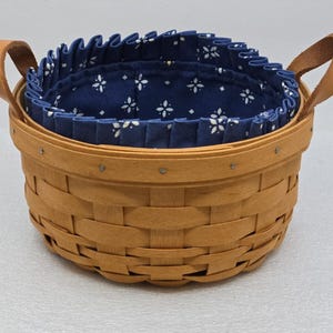 May include: A round woven wooden basket featuring a light brown finish and two tan leather handles attached with metal rivets. The interior is lined with a dark blue fabric featuring a white geometric star pattern and a pleated top edge.