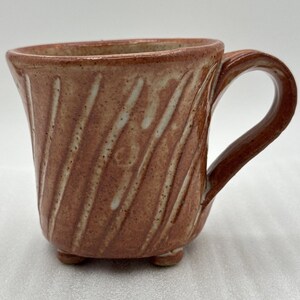 May include: A handmade ceramic mug with a reddish-brown glaze and a textured, diagonal stripe pattern. The mug has a curved handle and sits on three small feet. A unique piece for coffee or tea.