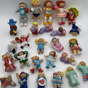 May include: A collection of twenty-five small Cabbage Patch Kids plastic figurines in various poses and outfits, including sports uniforms, casual clothing, and pyjamas, featuring diverse hair colours, skin tones, and expressions on a white surface.
