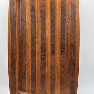 Puede incluir: Tabla de cortar rectangular de madera con ranura para jugos. La tabla está hecha de tiras verticales alternas de madera marrón clara y oscura. Las esquinas son redondeadas.