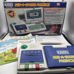 Puede incluir: Un juguete educativo electrónico VTech See-A-Sound Phonics en posición cerrada con carcasa azul y blanca, que incluye reproductor de casetes, botones de control y un asa. Se incluyen libros de ejercicios y la caja original del producto.