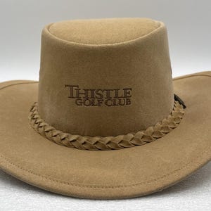 May include: A tan-colored hat with a wide brim and a flat-topped crown. The hat features a braided band around the base of the crown and the text "THISTLE GOLF CLUB" embroidered in dark brown thread on the front center of the crown.