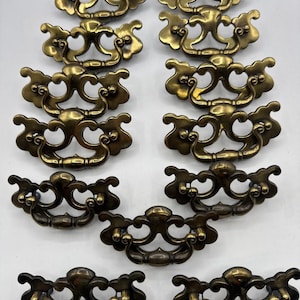 Puede incluir: Un conjunto de once tiradores de cajón de metal en tono latón, cada uno con una placa decorativa y ornamentada con volutas simétricas y un asa curva articulada. Los herrajes presentan un acabado pulido con un aspecto envejecido.