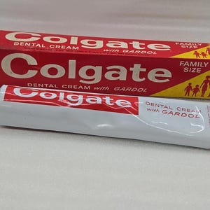 Pode incluir: Um tubo e uma caixa de pasta de dentes Colgate vintage. O tubo é branco com detalhes vermelhos e uma tampa vermelha. A caixa é vermelha com texto branco e uma seção amarela com uma silhueta familiar. O texto no tubo e na caixa diz "Colgate Dental Cream with Gardol."