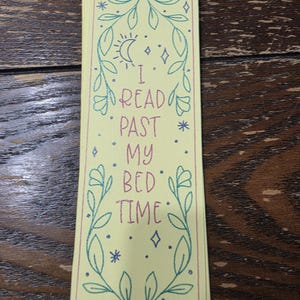 May include: A rectangular pale yellow bookmark featuring a decorative green leaf border, a crescent moon, and small stars. The text "I READ PAST MY BED TIME" is printed in the center in a pink font, framed by the botanical design elements.