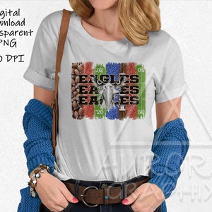 May include: A white t-shirt with a graphic design of a football field with the word "Eagles" repeated multiple times in black text. The graphic also includes a football and a stylized eagle head.