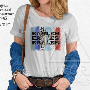 以下が含まれることがあります： イーグルの頭部が描かれたグラフィックデザインが施された白いTシャツ。グラフィックには「イーグル」という単語が3回繰り返されています。グラフィックは、ブルー、レッド、ホワイトの色で、使い古したような風合いになっています。