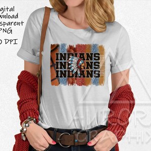 Könnte beinhalten: Ein weißes T-Shirt mit einem grafischen Design eines indianischen Kopfschmucks und dem Wort "INDIANS", das dreimal wiederholt wird. Die Grafik ist im Used-Look gehalten und in den Farben Rot, Orange und Blau gehalten.