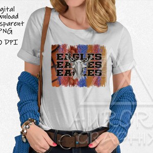 May include: White t-shirt featuring a graphic design of an eagle with the word "Eagles" repeated three times. The graphic is surrounded by a red, white, and blue design with basketballs.