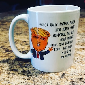May include: White ceramic coffee mug with a cartoon image of a person with orange hair and a red tie. The text on the mug reads "You're a really fantastic father. Great, really great. Wonderful, the best. Other fathers? Losers, total disasters! Terrible. Fake news. Believe me. Ask anyone."