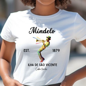 Puede incluir: Camiseta blanca con un diseño gráfico negro de una persona tocando la trompeta. El gráfico incluye el texto "Mindelo", "Est. 1879", "Ilha de São Vicente" y "Cabo Verde".