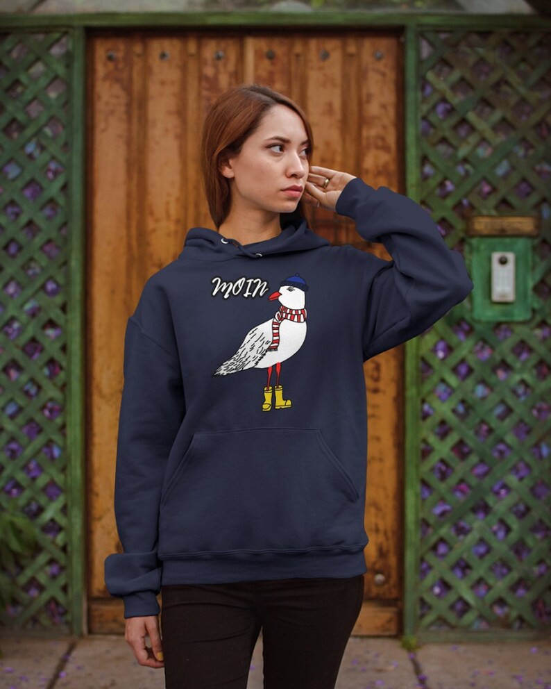 May include: A navy blue hoodie with a white seagull wearing a blue hat and red scarf. The seagull is wearing yellow rain boots. The hoodie has a pocket on the front and the word "Moin" printed on the front.