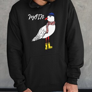 May include: Black hooded sweatshirt with a white seagull wearing a blue hat, red and white scarf, and yellow rain boots. The text "moin" is printed on the sweatshirt.